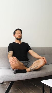 vertical view of a man sitting at the left side of a couch with his legs crossed and breathing deeply in a peaceful attitude