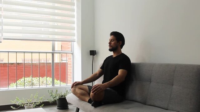 A hispanic man sitting on a couch with the legs crossed on a couch in a relaxing attitude breathing deeply in the morning