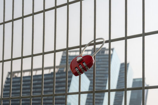 A single bright, red, heart-shaped lock with a combination is affixed to a fence on a bridge. Multiple tall skyscrapers are in the background.The love lock symbolizes commitment for two people in love