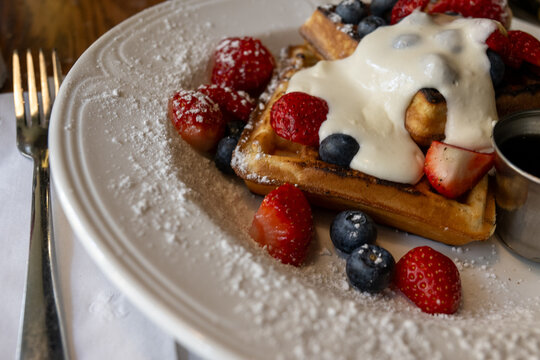 A round white plate with a square Belgian waffle topped with fresh cream and berries. The breakfast meal includes strawberries, blueberries, and raspberries. There's a small cup with maple syrup. A li