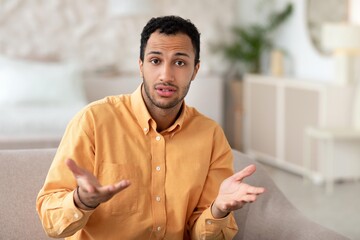 Worried Man With Short Hair Wearing Orange Shirt Expressing Confusion While Sitting On Sofa At...