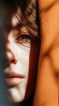 Close up portrait of a young woman with natural makeup, partially covered by an orange fabric, with dramatic sunlight and shadow patterns on her face. Artistic and emotional atmosphere