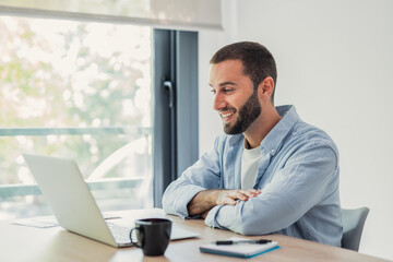 Fototapeta premium Man smiling while collaborating and communicating online through a laptop from his modern home office, symbolizing remote work, digital connectivity, and productivity