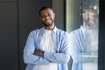 Fototapeta premium Confident young black man smiling, leaning against a modern window with his arms crossed, showing a happy expression reflecting success and positive emotions in an indoor setting