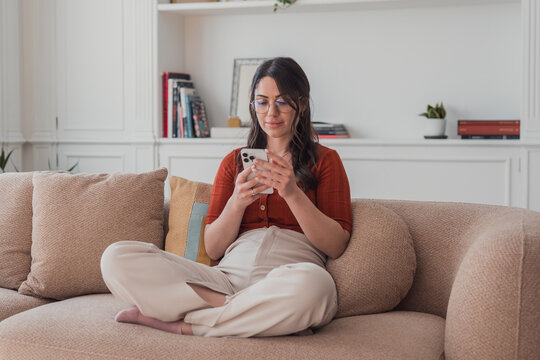 Woman sitting cross-legged on a modern sofa in her living room, smiling and engrossed in communicating or scrolling content on her mobile phone