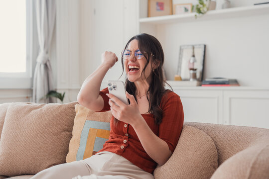 Young woman sitting on a sofa, looking at her smartphone and cheering with a fist pump, expressing excitement and joy after receiving good news or achieving success
