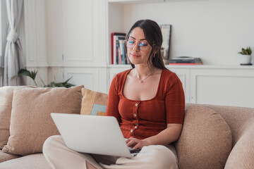 A young woman in glasses works on a laptop while sitting on a cozy couch in a bright living room, focused and relaxed as she types, researches and works remotely from home. © Daniel