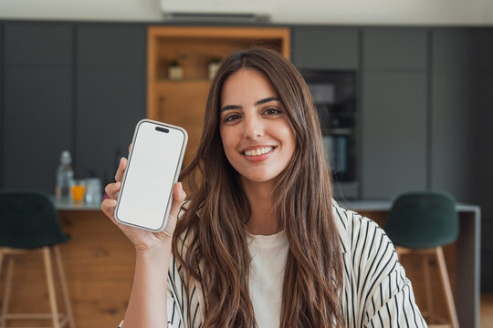 Smiling young woman sits in a modern kitchen holding a smartphone with a blank screen for app or website mockups, conveying friendliness, technology use, remote work and home lifestyle.