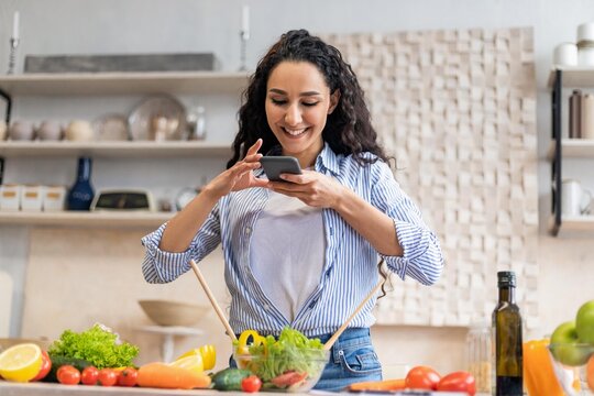 Smiling Young Woman Cooking And Taking Photo Of Ingredients While Preparing Fresh Salad In Modern Kitchen. Vibrant Atmosphere Reflecting Joy And Health, Emphasizing Healthy Eating Concept.