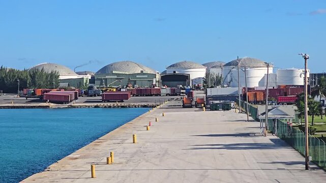 Wide shot of an empty concrete pier and dock leading towards a large industrial facility with storage domes, silos, and shipping containers at a commercial port on a sunny day.