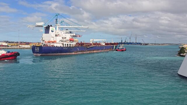 STI Singapore chemical tanker ship being maneuvering by two tugboats in a busy industrial container port, with blue container cranes in the background, under a partly cloudy sky and turquoise water.