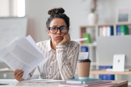 Worried Young Hispanic Woman Sitting At Desk In Office, Reading Documents With Concerned Expression, Exploring Important Information, Deep In Thought. Business Concept.
