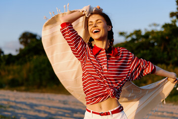 A young woman in a red striped shirt twirls on a sunlit beach, scarf fluttering behind her, hair loose, face lit with a bright, carefree smile, capturing a joyful lifestyle moment. © SHOTPRIME STUDIO