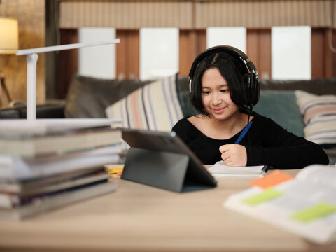 Asian teenage girl concentrates on online class, remote learning at home using tablet, writing and reading textbooks, self-study for back to school, Homeschooling, university education lifestyle.
