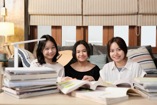 Three Asian teenage girls, friends, are cheerfully self-learning tutorials at home together, reading textbooks, preparing school homework exams, Homeschooling study, smiling, and looking at camera.