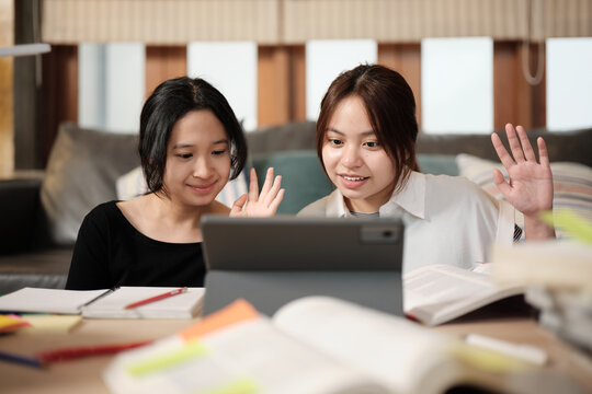 Two Asian teenage girls greeting in online class, remote learning at home using digital tablet, writing and reading textbooks, self-study for back to school, homeschooling education knowledge.