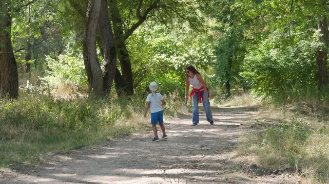 A loving mother bends down while playing badminton with her happy little boy. They share a joyful moment on a dirt path in a green park.