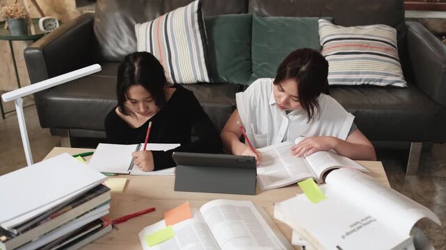 Two Asian teenage girls concentrate on online class, remote learning at home using tablet, writing and reading textbooks, self-study for back to school, Homeschooling, university education lifestyle.