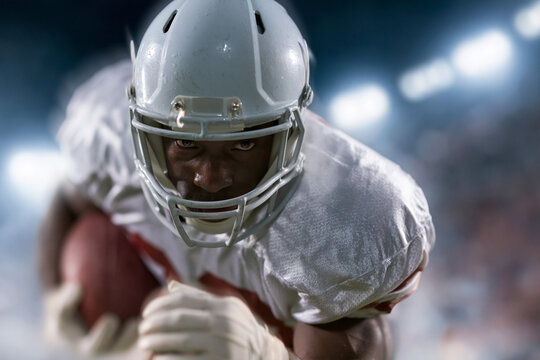 Football player grips ball tightly, eyes focused forward. Helmet gleams under stadium lights, fierce determination visible. Dense crowd blurred behind him, spotlight intensifies his resolve