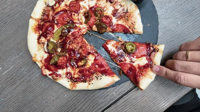 Freshly baked pepperoni and jalape&ntilde;o pizza viewed in a close-up shot. A hand lifts a cheesy slice from a round slate plate, showing melted cheese and vibrant toppings in natural daylight.
