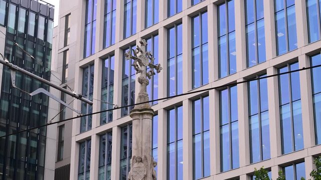 St Peter's Cross stands in front of glass and stone office buildings with tram wires cutting across the frame in Manchester city centre.