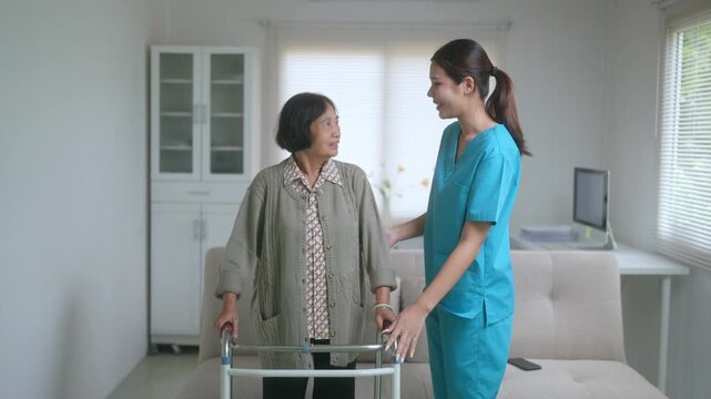 Young Asian nurse supporting an elderly woman during rehabilitation at home, guiding her while she uses a walker for mobility and physical therapy exercises in the living room