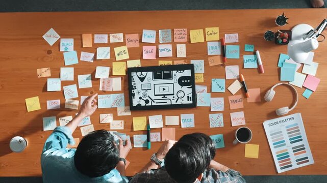 Top view of creative designer working together brainstorming idea by using sticky notes at meeting table with color palettes. Aerial view of smart team choosing icon graphic from tablet. Symposium.