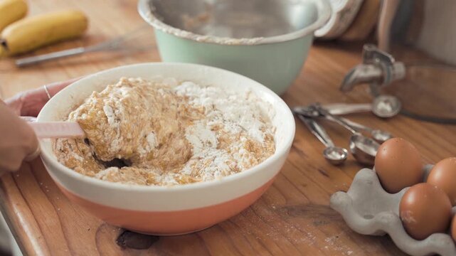 Baker mixes fresh banana bread batter in a ceramic bowl using a silicone spatula while wooden kitchen countertops provide a warm, rustic background setting for this classic home culinary baking.