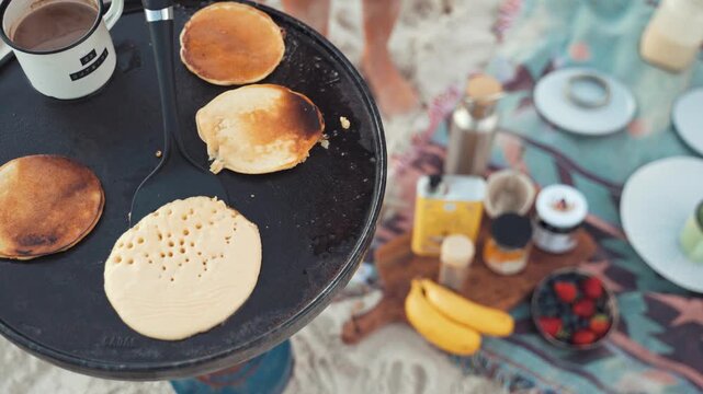 Pancakes cook on a black iron griddle outdoors while a morning picnic spread sits on a blanket nearby, offering a rustic breakfast experience during a relaxed camping trip in the bright natural light.