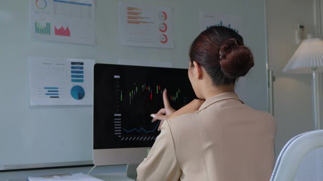 Back view of a young asian woman analyzing financial candlestick charts for stock trading, monitoring market trends and making investment decisions while working late in the office