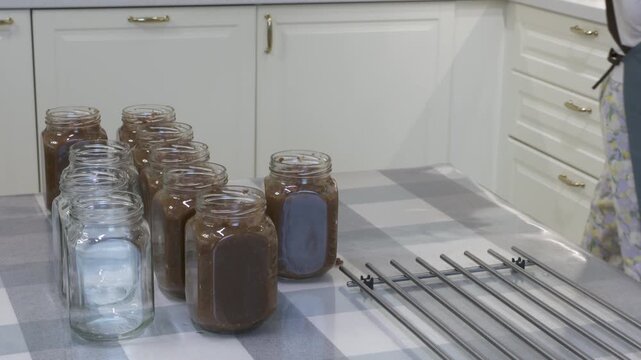 Glass jars filled with homemade preserves line a table in a bright kitchen. The neat arrangement feels satisfying and calm. The warm room brings cozy comfort.