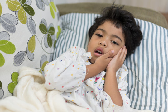 Unwell preschooler girl lying on sofa touching her cheek in pain. Suffering from severe toothache or mumps illness, highlighting urgent pediatric dental care and treatment