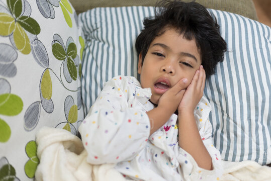 Exhausted little girl suffering from toothache pain while resting on a sofa. Exhibiting pediatric dental problems or viral mumps illness requiring immediate medical care