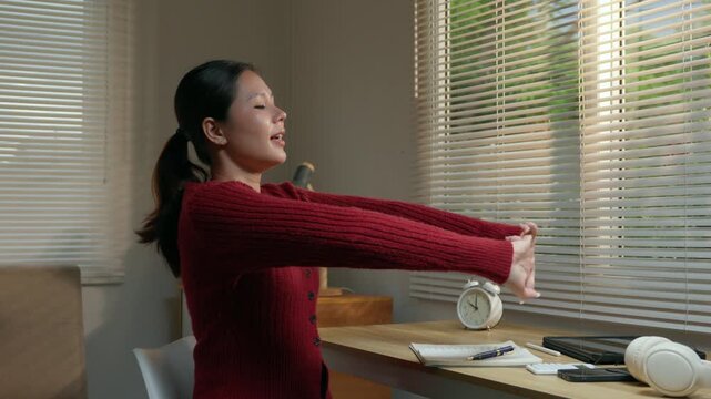 Relaxed young asian woman sitting at her desk, stretching her arms and back after finishing work, feeling happy and relieved while enjoying a break in her cozy home office