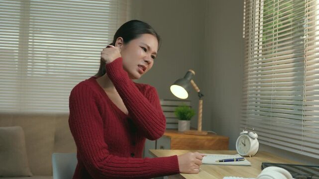 Overworked asian woman massaging her tense neck and shoulder muscles while sitting at her desk, experiencing pain and discomfort due to poor posture and long hours of sedentary work