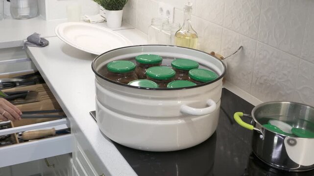 Glass jars with green lids warm inside a large white pot on a stovetop. A woman reaches into an open drawer in a clean kitchen. The scene feels peaceful and cozy.