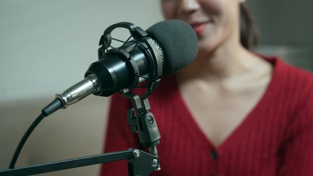 Young asian woman speaking into a professional condenser microphone while hosting a live radio show or recording a podcast in a home studio, with a focus on the audio equipment