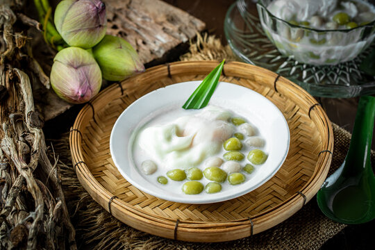 Thai dessert Bua Loi with pandan rice balls and sweet egg in coconut milk, served in a rustic zinc bowl on bamboo tray with pink lotus flowers and vintage clay pot background