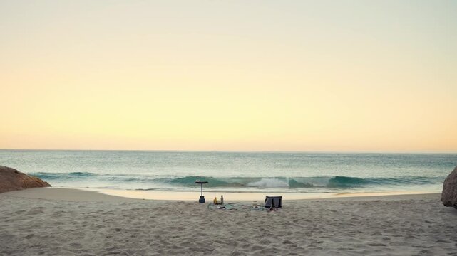A serene beach landscape features gentle waves rolling onto a sandy shore under a soft, warm sky, with a few scattered items suggesting a peaceful outdoor picnic setting during golden hour.