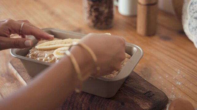 Hands adorned with gold bracelets are carefully arranging sliced bananas on top of raw cake batter inside a metal loaf pan on a wooden table to ensure a beautiful finish before the baking process.