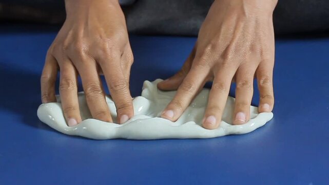 human hands pressing and flattening soft white stretchy slime onto a plain blue surface.