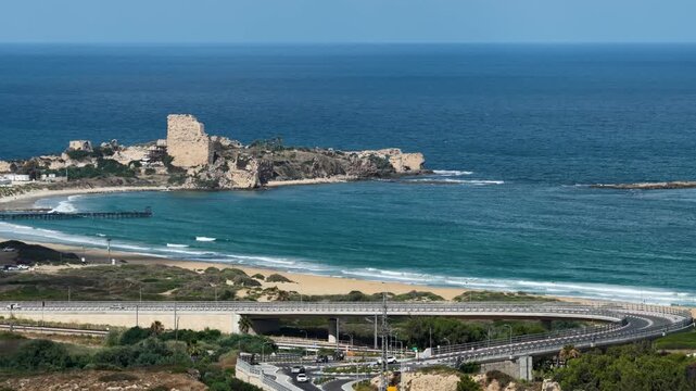 High angle shot showcasing the contrast between the historical Chateau Pelerin crusader castle ruins on the coastline and a contemporary curved highway bridge with active traffic under a blue sky