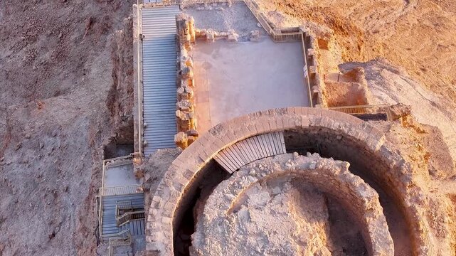 Drone pull back shot capturing a top down perspective of the ancient circular stone tower and viewing deck at Masada fortress with the Judean desert ground textures expanding behind.