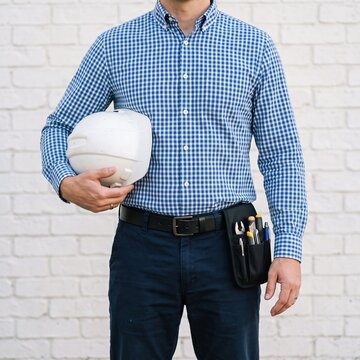 Man in a checkered blue shirt holds a white safety helmet under his arm, wearing a compact tool pouch on his belt against a white brick wall.