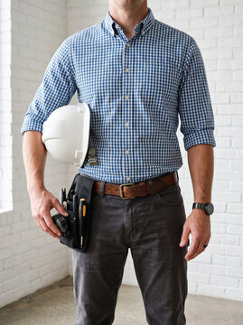Man in a checkered blue shirt holds a white safety helmet under his arm, wearing a compact tool pouch on his belt against a white brick wall.