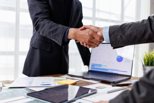 Business partners shaking hands, symbolizing successful collaboration, agreement, partnership, and a strong deal closure after analyzing financial data and charts on a laptop