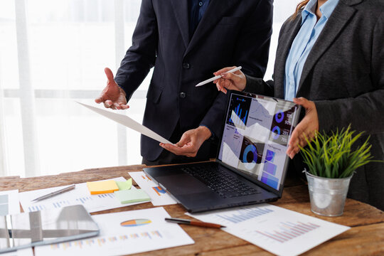 Business professionals collaborating at a wooden desk. Reviewing financial reports and digital dashboards on a laptop. Exchanging ideas