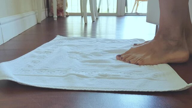 Barefoot woman steps onto a white towel placed on wooden floor, showcasing foot placement and texture in a bright indoor setting with natural light