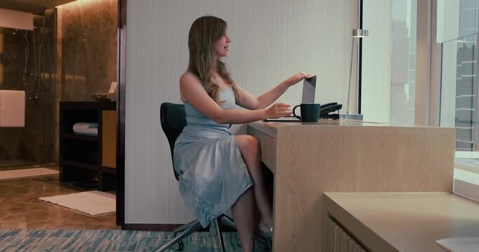 Young Caucasian woman in blue dress finishes work, closes laptop, and picks up black coffee mug at a desk in a modern hotel room. City view through window. Medium shot of business traveler.