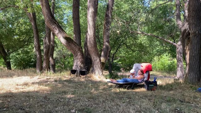 A loving mother and her small son relax together on a lounge chair. They rest peacefully under large trees in a sunny forest. This cozy moment brings pure joy.